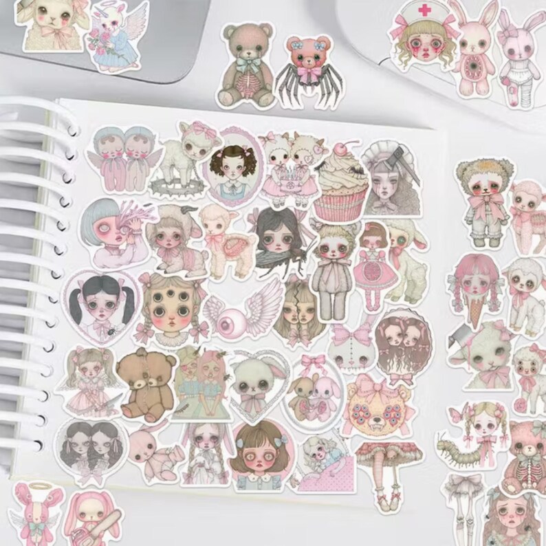 May include: A collection of stickers featuring various illustrations in a pastel color palette. The stickers depict dolls, teddy bears, bunnies, and other whimsical characters. The stickers are arranged on a white spiral-bound notebook.