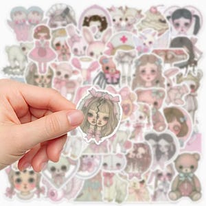 May include: A collection of pastel-colored stickers featuring various illustrations. The stickers include images of dolls, bunnies, teddy bears, and other cute characters. One sticker is held in a hand, showcasing two doll faces with pink bows.