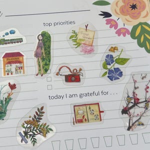 May include: A planner page featuring decorative stickers. Stickers include a train, a flower shop, a girl, a camera, and floral designs. The text "top priorities" and "today I am grateful for..." are visible.