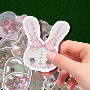 May include: A white bunny sticker with a pink bow and stitched details is held in a hand. The bunny has large eyes and a stitched mouth. Other stickers with similar themes are in the background.