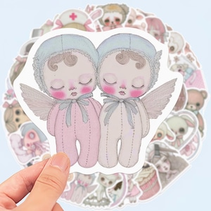 May include: A hand holding a sticker sheet with multiple stickers. The primary sticker features two baby dolls with closed eyes, wings, and bonnets. The dolls are wearing pink and cream-colored onesies. Other stickers include teddy bears and bunnies.