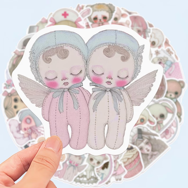 May include: A hand holding a sticker sheet with multiple stickers. The primary sticker features two baby dolls with closed eyes, wings, and bonnets. The dolls are wearing pink and cream-colored onesies. Other stickers include teddy bears and bunnies.