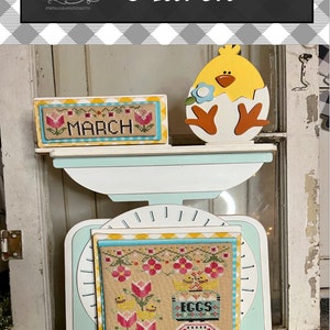 May include: A white and blue vintage scale with a yellow chick figurine and two cross-stitch pieces. The cross-stitch pieces are framed in yellow and blue and feature a floral design with the words "March" and "Eggs".
