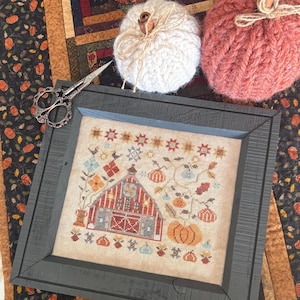 May include: A framed cross-stitch featuring a barn with pumpkins and fall foliage. Two knitted pumpkins, one white and one coral, sit nearby. A pair of decorative scissors rests on the frame. The scene is set on a fall-themed runner.