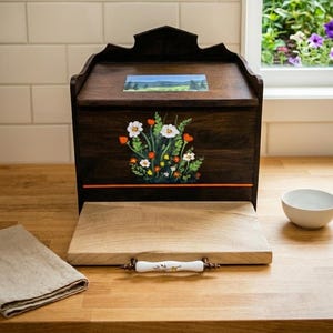 Peut inclure: Une boîte à pain en bois brun foncé avec une porte frontale à charnières ornée d'une composition florale peinte à la main de fleurs blanches et orange. Le couvercle présente un petit paysage peint et la porte inclut une poignée en céramique.