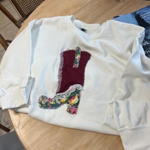 May include: White sweatshirt with a red, white, and floral boot design appliqued on the front.