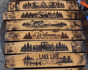 May include: A collection of eight curved wooden barrel staves featuring laser-engraved designs including hunting gear, a campfire, a camping scene, a jumping fish, text reading Welcome to our neck of the woods, a truck, Lake Life, and Dad's Garage.