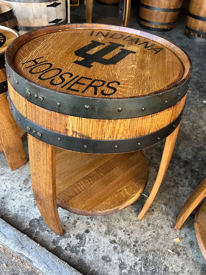 May include: A wooden barrel end table with a black metal band and a top shelf. The table is decorated with the Indiana Hoosiers logo and the words "Indiana Hoosiers".
