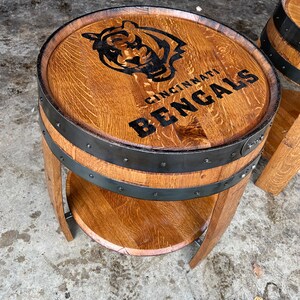 May include: A wooden barrel end table with a black metal band and a shelf. The top of the table is decorated with a black and white logo that says "Cincinnati Bengals".