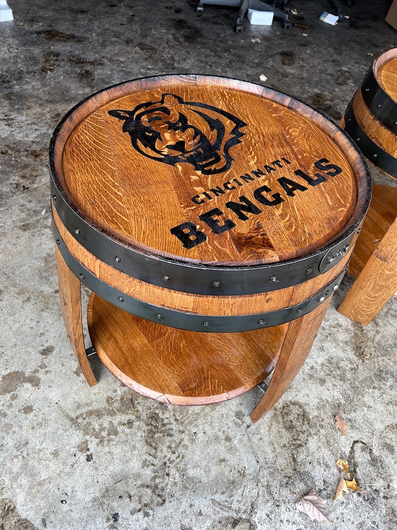May include: A wooden barrel end table with a black metal band and a shelf. The top of the table is decorated with a black and white logo that says "Cincinnati Bengals".