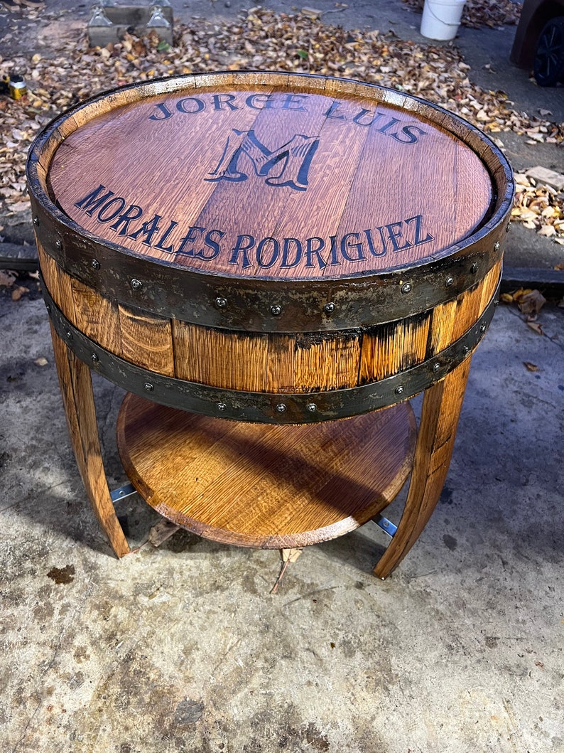 May include: A wooden barrel end table with a metal band and a shelf. The top of the table is engraved with the text "Jorge Luis Morales Rodriguez" and a large "M".