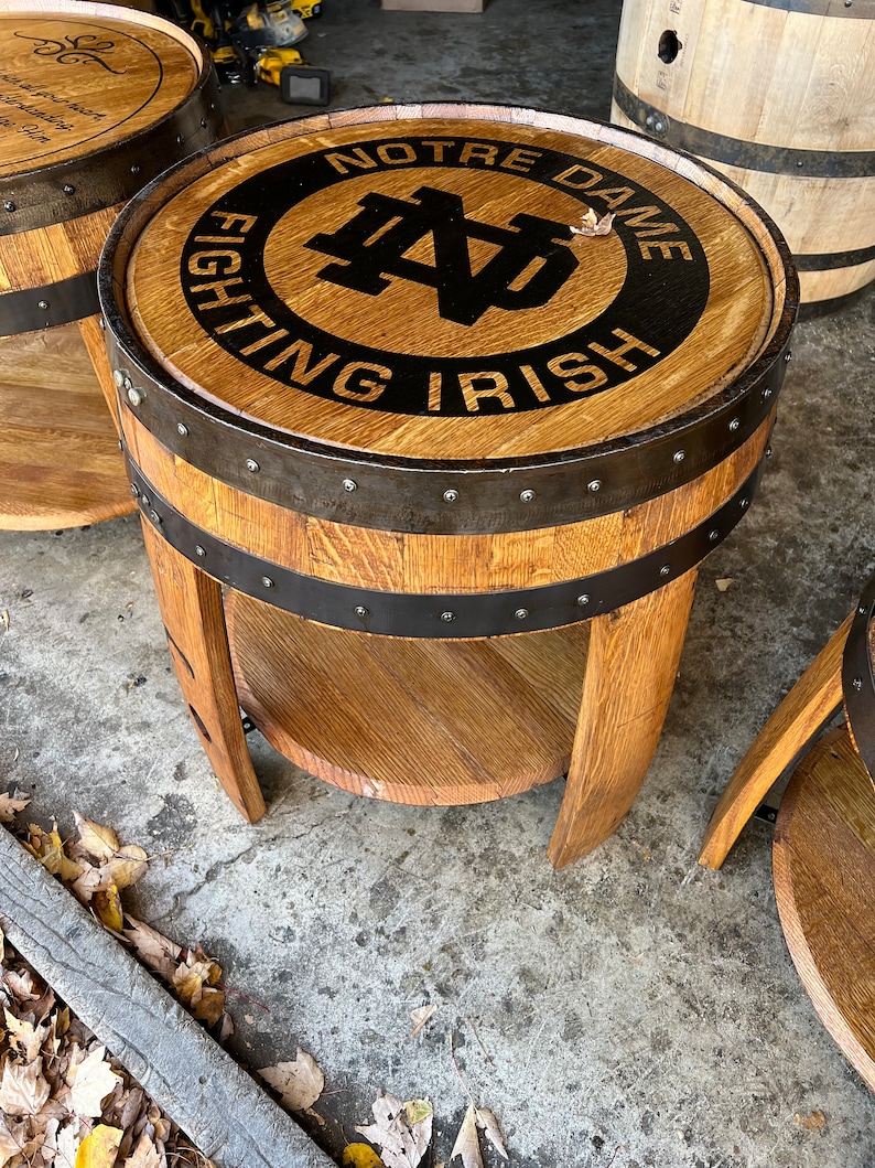 May include: A wooden barrel end table with a black and gold Notre Dame Fighting Irish logo. The table has a shelf and metal bands around the top and bottom.
