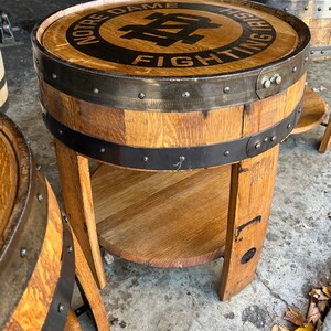 May include: A wooden barrel end table with a black metal band and a circular top featuring the Notre Dame Fighting Irish logo. The table has a lower shelf and is made from reclaimed wood.