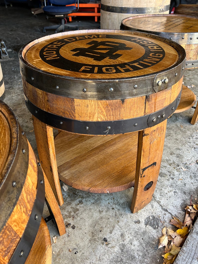 May include: A wooden barrel end table with a black metal band and a circular top featuring the Notre Dame Fighting Irish logo. The table has a lower shelf and is made from reclaimed wood.