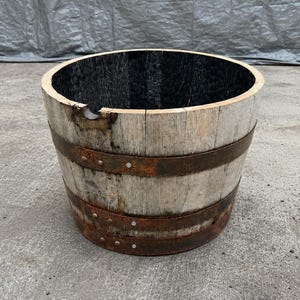 May include: A cylindrical wooden barrel planter featuring weathered light-colored oak staves held together by two wide, rusted metal bands. The interior of the barrel is charred black, and there is a small notch cut into the top rim of the wood.