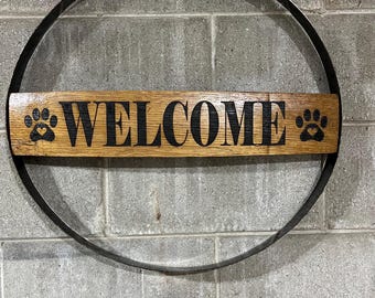 May include: A wooden sign with the word "WELCOME" in black lettering and two paw prints on either side, mounted on a black metal ring.