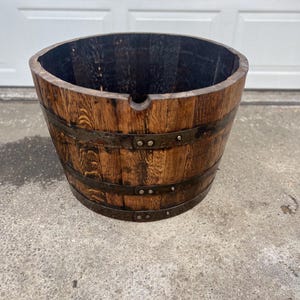May include: A wooden barrel planter with dark brown wood and metal bands. The barrel has a round shape and a dark interior. The metal bands are secured with rivets. The planter is sitting on a concrete surface.