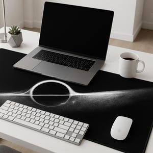 May include: A black desk mat with a white graphic design, a laptop, a white keyboard, a white mouse, a white mug, and a black smartphone on a white desk. A small potted plant is also visible.