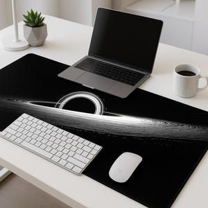 May include: A black desk mat featuring a white and gray graphic of a black hole. A laptop, white keyboard, mouse, and coffee mug are on the mat. A smartphone and small potted plant are also on the white desk.