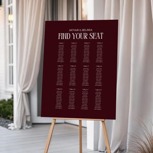 Könnte beinhalten: Ein weinroter Sitzplan auf einer Holzstaffelei. Die Tafel zeigt den Text "ARTHUR & BELINDA FIND YOUR SEAT" in Weiß, mit den Namen der Gäste nach Tischnummer geordnet. Die Staffelei ist aus hellem Holz gefertigt.