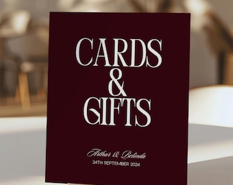 May include: A rectangular dark burgundy sign with white text that reads CARDS & GIFTS, followed by the names Arthur & Belinda and the date 24TH SEPTEMBER 2024. The sign is supported by a thin, horizontal gold-colored metal stand base.