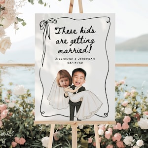 May include: A white wedding sign featuring a black hand-drawn border with a bow. The text reads "These kids are getting married!" followed by "JILLIANNE & JEREMIAH" and "08/14/28". It includes a cartoon illustration of a child bride and groom.