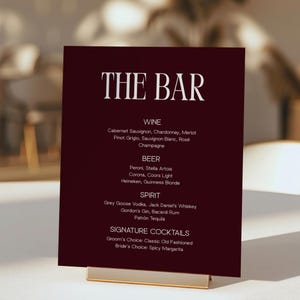 May include: A rectangular dark burgundy bar menu sign with white text reading THE BAR, WINE, BEER, SPIRIT, and SIGNATURE COCKTAILS, listing various drink options including Cabernet Sauvignon, Peroni, Grey Goose, and Old Fashioned, held by a gold metal stand.