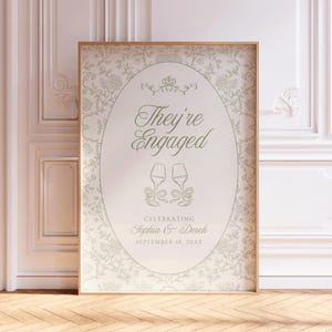 May include: A rectangular framed sign featuring a light beige background with a gray floral pattern. The center oval contains the text "They're Engaged" above two wine glasses with bows, followed by "Celebrating Sophia & Dereh" and "September 18, 20XX".