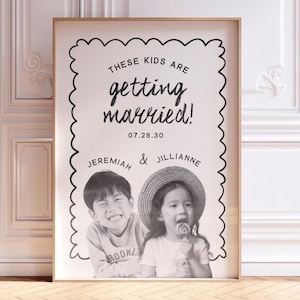 Op de afbeelding: Een ingelijste verticale poster met een geschulpte zwarte rand op een witte achtergrond. De tekst luidt: THESE KIDS ARE getting married! 07.28.30 JEREMIAH & JILLIANNE. Onder de tekst staat een zwart-witfoto van twee lachende kinderen.
