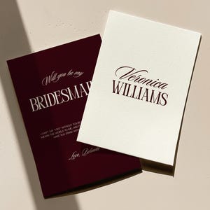 May include: Two rectangular cards. The top card is cream-colored with the dark maroon text 'Veronica WILLIAMS' in a mix of script and serif fonts. The bottom card is dark maroon with white text reading 'Will you be my BRIDESMAID'.