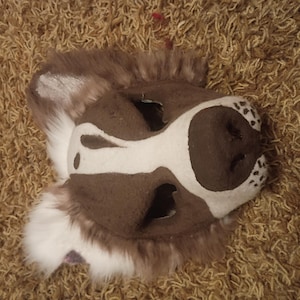 May include: A brown and white animal mask featuring faux fur trim, pointed ears, and a snout with black spotted markings. The mask has two eye openings and a central white stripe extending from the forehead down to the nose area.