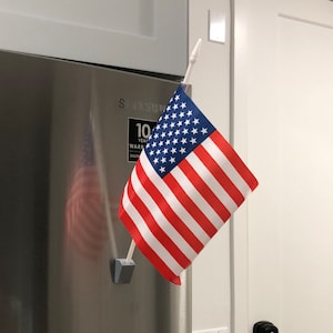 May include: A small American flag attached to a refrigerator with a magnetic flag holder. The flag is red, white, and blue with stars on the blue field.