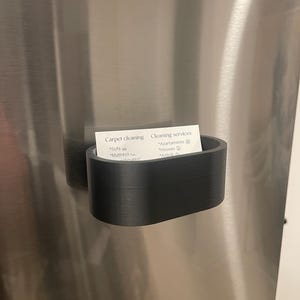 May include: A black, curved business card holder attached to a stainless steel surface. The holder contains a white card with text about carpet cleaning and cleaning services.