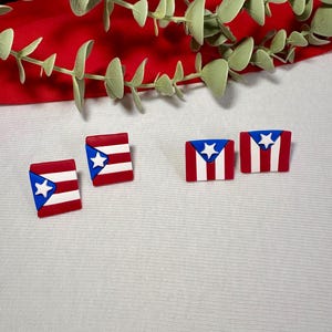 May include: Four small, square-shaped accessories featuring the Puerto Rican flag design. Each piece has a white star on a blue background and red and white stripes. The accessories are displayed on a white surface with a red background and green leaves.