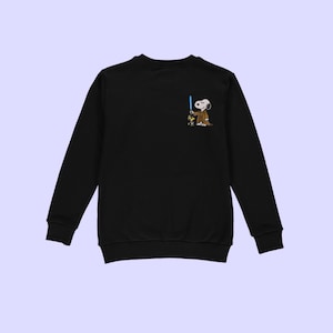Puede incluir: Sudadera negra de manga larga con un personaje de dibujos animados de Snoopy vestido de Jedi, sosteniendo un sable de luz azul. El gr&aacute;fico est&aacute; en la parte superior izquierda del pecho. La sudadera tiene cuello redondo y pu&ntilde;os acanalados.