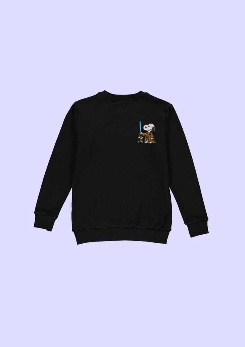 Puede incluir: Sudadera negra de manga larga con un personaje de dibujos animados de Snoopy vestido de Jedi, sosteniendo un sable de luz azul. El gr&aacute;fico est&aacute; en la parte superior izquierda del pecho. La sudadera tiene cuello redondo y pu&ntilde;os acanalados.