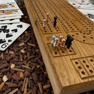 Può includere: Un tabellone di cribbage in legno con finitura naturale, con file di fori praticati e aree di punteggio scolpite. Il tabellone include pioli argentati, ramati e neri. Le carte da gioco sono visibili sullo sfondo.