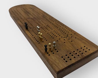 May include: A wooden cribbage board with a dark brown finish. The board has multiple rows of small holes and includes a set of metal pegs in various colors, including black, silver, and gold. The board is rectangular with rounded ends.