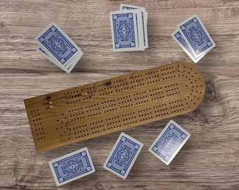 May include: A wooden cribbage board with a rounded end and numerous holes, surrounded by playing cards with blue and white designs. The board has metal pegs and is set on a wooden surface.