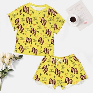 May include: A bright yellow pajama set consisting of a short-sleeved henley top and matching shorts. The fabric features a repeating pattern of two women's faces, green hearts, red hearts, and the text Lucy & Sophia printed across the entire surface.