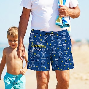 May include: Navy blue swim trunks with a pattern of stick figures, stars, and the text "This Grandpa Belongs to" followed by names. The trunks have a drawstring and are worn by a person on a beach.