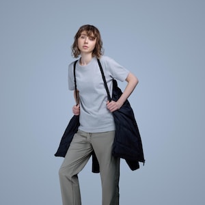 May include: A person wearing a light grey t-shirt and khaki trousers, with a black quilted jacket draped over the shoulders. The jacket has black straps. The background is a solid light blue.