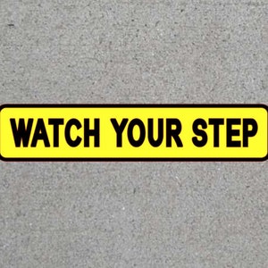May include: A rectangular yellow sign with black text that reads "WATCH YOUR STEP." The sign has a black border and is set against a gray concrete background. The sign is designed to alert people to be cautious.