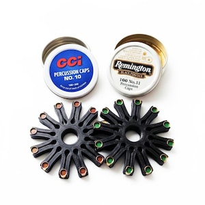 May include: Two black plastic circular percussion cap dispensers with radial arms holding metallic caps, positioned below a blue CCI No. 10 percussion caps tin and a white Remington No. 11 percussion caps tin with black and gold text.