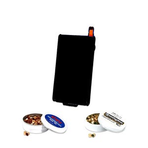 Puede incluir: Un dispensador rectangular negro de fulminantes con punta naranja, acompañado de dos latas metálicas redondas. Una lata tiene la etiqueta CCI Percussion Caps No. 10 con fulminantes de cobre, y la otra tiene la etiqueta Remington No. 11.
