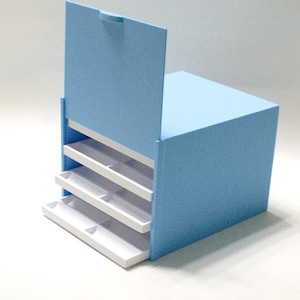May include: A light blue storage organizer with a vertical flip-top lid and three white pull-out drawers. The exterior of the unit has a textured finish, while the interior drawers are smooth and white, providing a contrast to the blue frame.