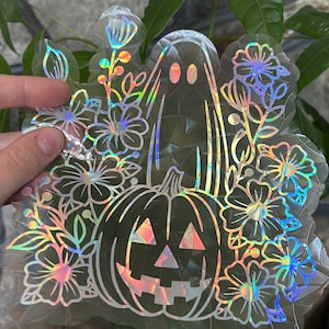 May include: A holographic sticker featuring a pumpkin with a spooky face, a ghost, and floral designs. The sticker has a rainbow iridescent finish.