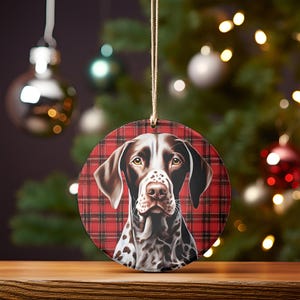 Puede incluir: Un adorno navideño redondo con un perro Braco Alemán de Pelo Corto marrón y blanco sobre un fondo de cuadros rojos y negros.