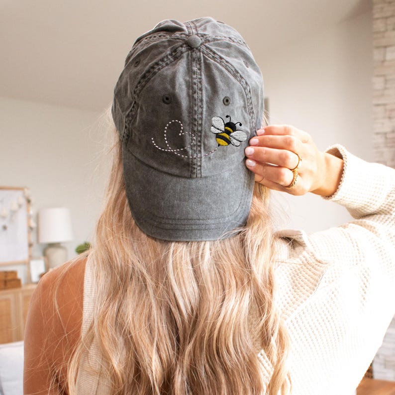 Puede incluir: Gorra de b&eacute;isbol gris con un acabado desgastado. La gorra presenta un dise&ntilde;o bordado de una abeja amarilla y negra con una trayectoria de vuelo punteada. La gorra es un dise&ntilde;o cl&aacute;sico de seis paneles con una visera curva.