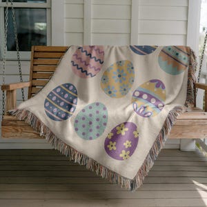 May include: A cream-colored woven throw blanket with a fringe border features a pattern of colorful Easter eggs. The eggs are decorated with various designs, including polka dots, stripes, and floral patterns.